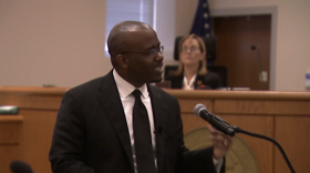 A man in a black suit and tie speaks into a microphone in a courtroom, with a judge seated in the background.