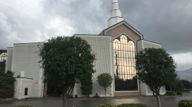 A white church with a tall steeple and mirrored glass on the front surrounded by several trees