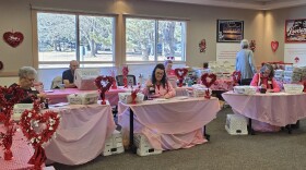 People sit at six tables, covered with pink tablecloths and adorned with shiny red heart decorations. They are stamping envelopes with a special heart symbol for the Loveland Valentine Remailing Program.