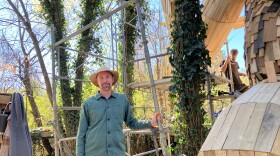 A wood sculptor stands outdoors next to his wooden statue.