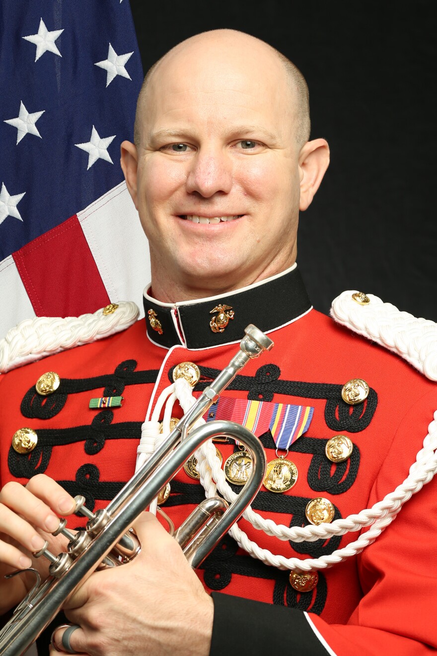 A man in a red military-style jacket sits in front of a U.S. flag and holds a trumpet in this portrait.