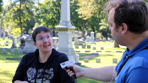 A man with a microphone interviews a woman in a cemetery. 