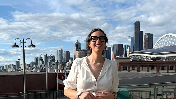 A smiling woman on a sunny day standing on a peer with the Seattle city scape in the background. She is wearing a white dress and dark rimmed glasses. 