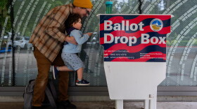 FILE - Matt lifts up his three-year-old son, Theo, to cast his ballot into the Hillsboro Public Library-Shute Park ballot drop box in May 2026. Election officials are increasingly recommending voters plan to use drop boxes rather than mailing their ballot.