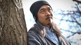 A man with a stocking cap and long hair wears a light jacket and is leaning against a tree looking off camera. He's got a slight smile on his face.