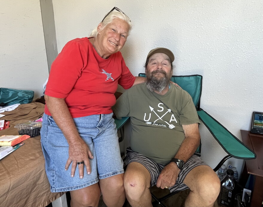 A middle-aged man and woman sit with their arms around each other. She has white hair and he has a long gray beard. 