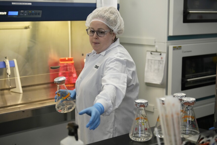 Operational technician Erin Aranda, wearing a white lab coat and hair net, holds a glass flask in her right hand in an industrial facility. 