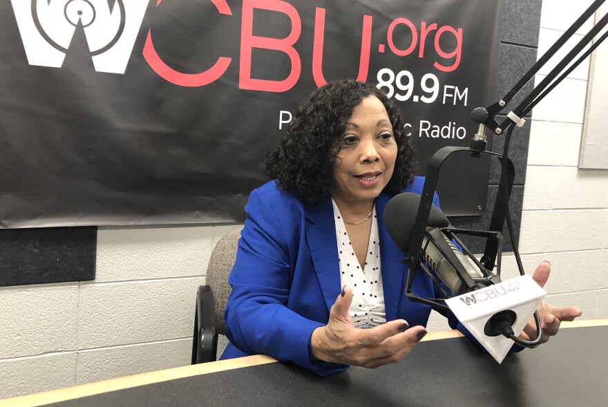 A woman in a blue jacket and white dotted blouse speaks into a microphone as she sits at a table in the WCBU master studio, with the station's branding logo banner mounted on a white concrete wall behind her. 