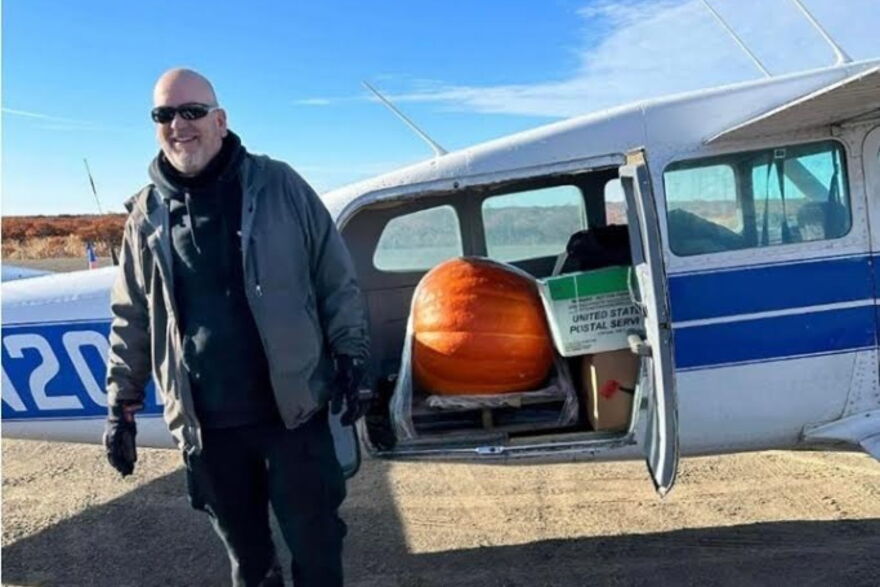 The winner of a pumpkin weight guessing contest hosted by an AC grocery store smiles outside of a bush plane with his prize waiting inside.