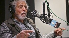 A man inside a radio studio sits at a microphone. He is pointing with both hands while talking.