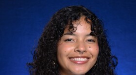 Malory Shaw with long and curly, dark hair. She is standing in front of a blue backdrop and smiling.
