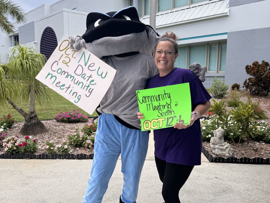 A person inside of a blue and gray stingray mascot costume stands holding a white sign, while the other arm is around the shoulder of a woman wearing a purple shirt and black leggings, holding a green sign. Both signs note a community meeting taking place on Oct. 12.
