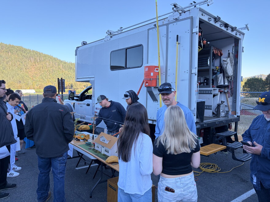 Students and presenters surround a tabletop model of power lines beside a utility truck at an outdoor career fair.