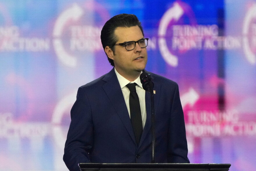 A man in a dark blue suit with white shirt and black tie stands at a lecturn or podium. He's wearing glasses and speaking into a mic.