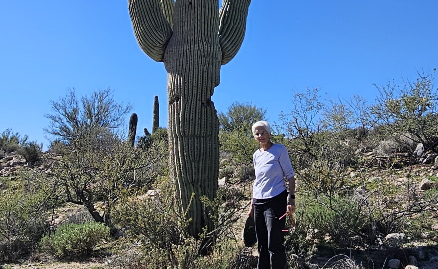 older woman with white hair stands next to monstrous saguaro cactus in the middle of a desert landscape