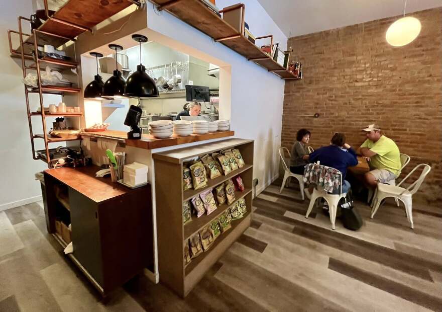 A cozy cafe interior with shelves of snacks and plates near a kitchen window. Three people sit around a table by a brick wall, having a conversation. The space has wood accents and modern lighting.