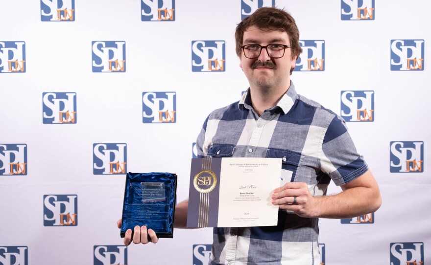 Reporter Clayton Baumgarth standing in front of a white backdrop with the SPJ logo. He is a white man wearing a plaid shirt and glasses, holding a glass award and paper certificate.