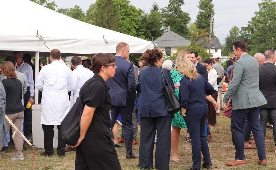 A bustling outdoor event with people in formal and casual attire gathered under tents. Trees and a building are visible in the background.