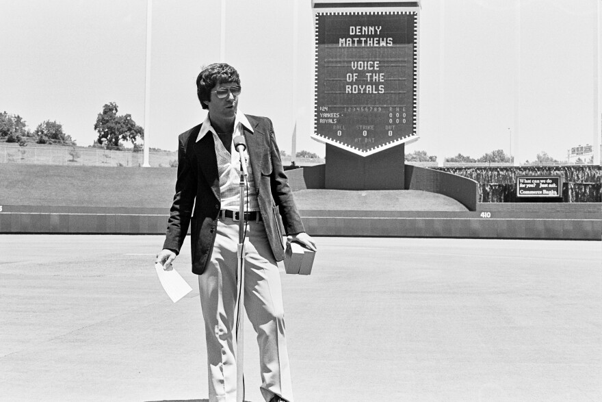 A young Denny Matthews stands on the diamond at the K wearing aviators. Behind him on the crown board has the words "DENNY MATTHEWS -- VOICE OF THE ROYALS."