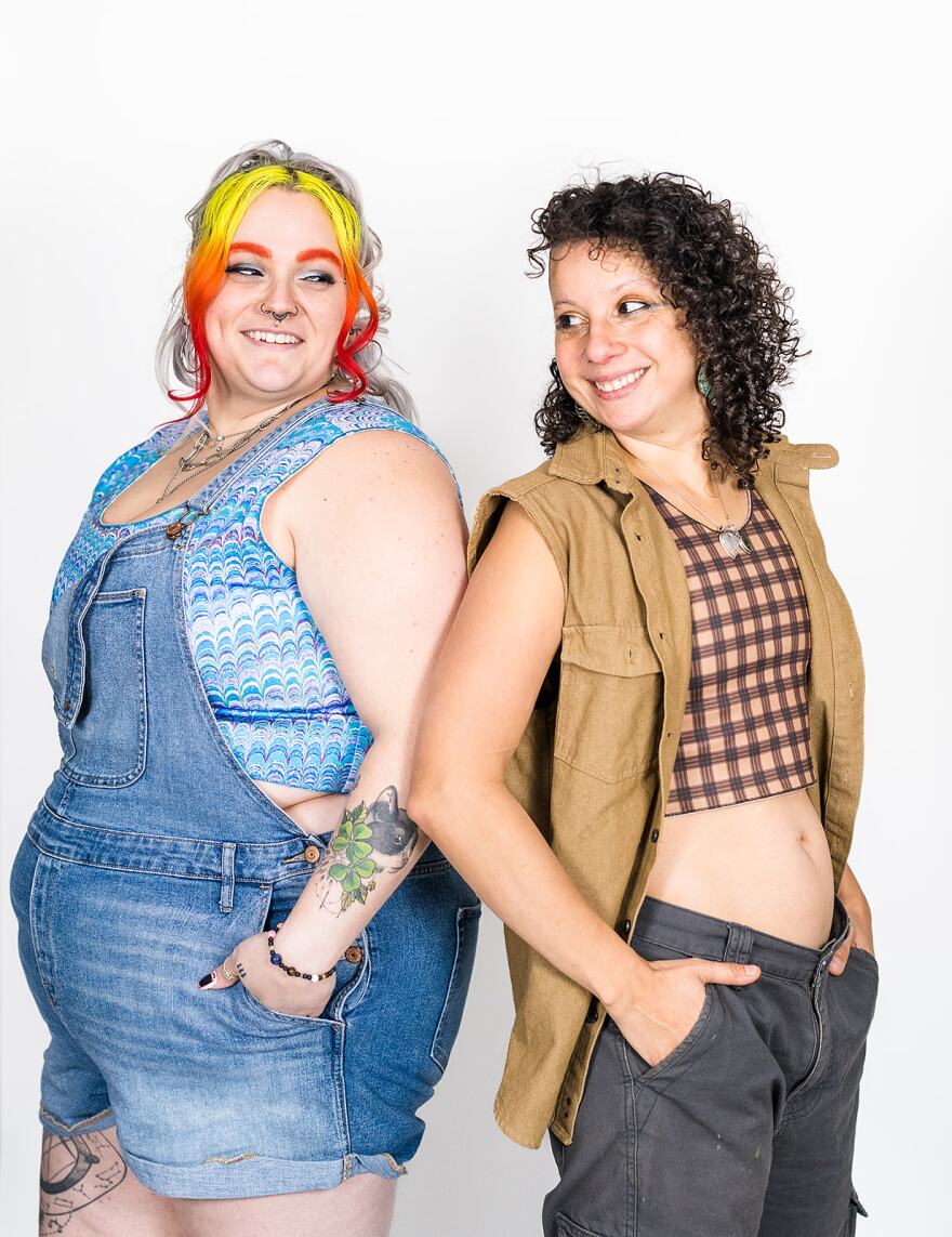 One model wearing overalls and a blue binder with another model wearing grey cargo pants, brown binder and sleeveless jacket. They are back-to-back and smiling.