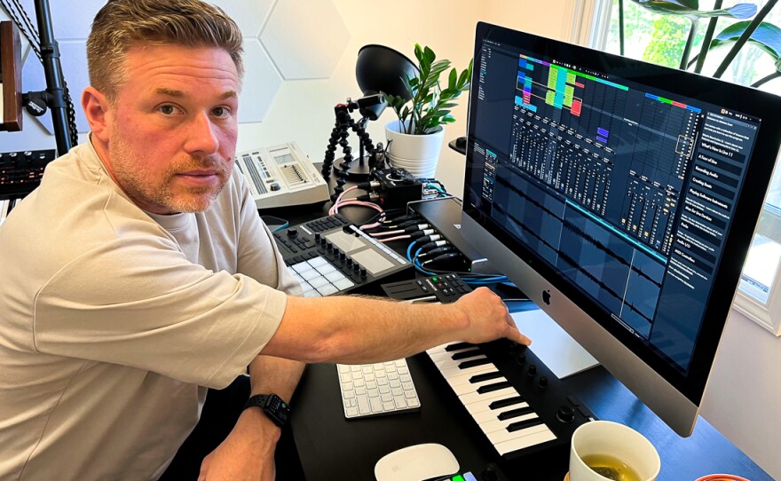 A man looks at the camera with his hands on a small piano keyboard, in front of a computer screen.