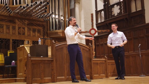 Pete Buttigieg speaks into a microphone and points at Sean McCann, who is standing next to him, on a wooden church stage in front of a large pipe organ. Both are white men with dark hair, and both are wearing light button-up shirts and dark pants. McCann is looking at Buttigieg and smiling. 