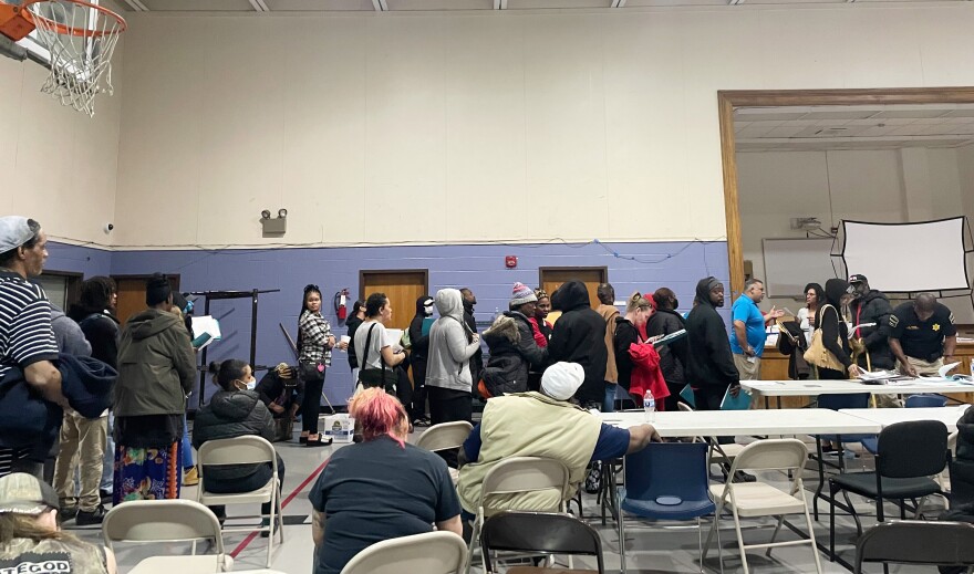 Indoor photo inside a gymnasium. People are standing in line. There are lots of metal folding chairs and tables in the foreground. Some people are sitting. Most are standing.