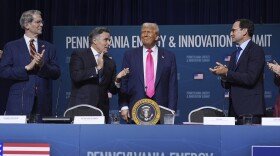 A white man in a suit smiles while standing behind a table with the presidential seal on it as three other white men look at him and applaud.