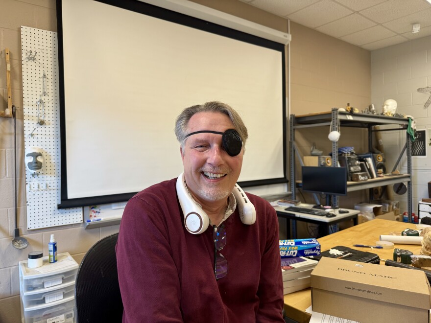 A man posses for a photo in the art studio with an eye patch and neck fan.