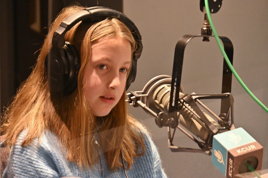 A young girl wearing a blue sweater sits behind a microphone in a studio setting.