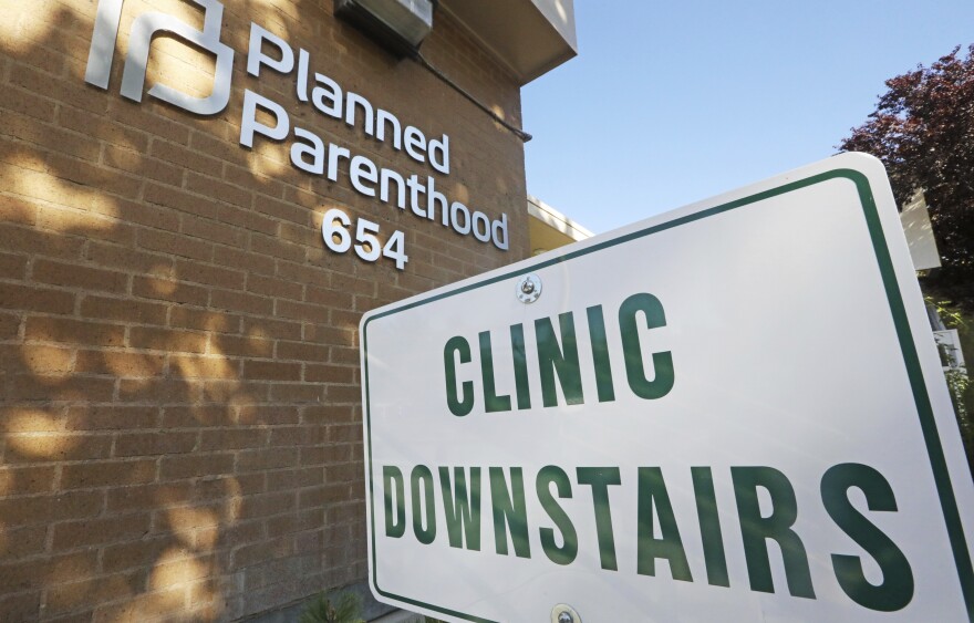 The words Planned Parenthood with the Planned Parenthood logo on a brick building and a white sign in front that says Clinic Downstairs. 