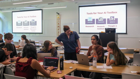 High school students with laptops gather around a table. A college student and an adult have a conversation. Behind them, projector screens read "Tools for Your AI Toolbox: Research, Research Assistant, Thought & Brainstorm Partner." 