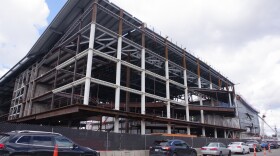 A line of cars drives past a large, metal structure with exposed beams. Workers walk along the structure. and machines can be seen in the distance. It is an active construction site. 