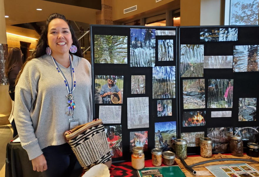 Teara Farrow Ferman stands in front of a display of photographs and containers documenting and displaying first foods. Farrow Ferman wears large, colorful, circular beaded earrings, she has long, dark hair and is smiling.