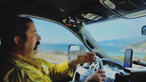 A man in a yellow firefighting jacket drives on a highway with the ocean behind him. 