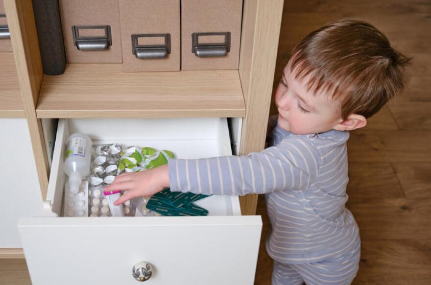 Toddler baby opened the cabinet drawer with pills and medicine. Child boy holding a pack of pills in the home living room. Kid age one year nine months