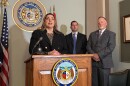 A woman in a black blazer and leopard print shirt speak at a podium with a microphone. The front of the podium has the seal of the state of Missouri on it. Two men in suits stand behind her. 
