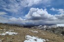 This is an image taken from a mountain peak above Reno. The land is mostly brown with small patches of white snow. The sky is blue with white puffy clouds. 