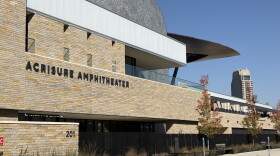 A view of the sandstone-colored brick facade of the Acrisure Amphitheater in downtown Grand Rapids. To the right of the frame, you see the roof of the amphitheater, open to the city, with a high-rise in the distance.