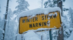 Yellow winter storm warning sign covered in snow and icicles in a cold forest setting. Ideal for themes of extreme weather, travel safety and wilderness conditions.
