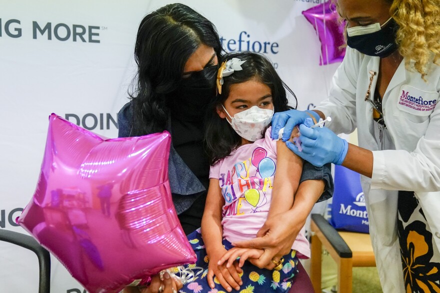 A young girl sits on a woman's lap and receives a shot in her left arm from a nurse. All three wear masks, the girl has a ribbon in her hair and holds a star shaped balloon.  