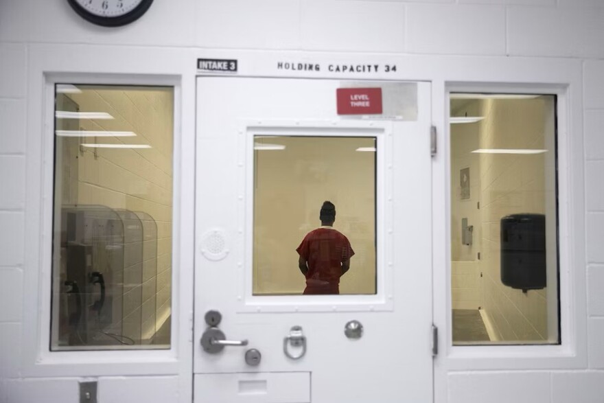 A person in a prison jumpsuit stands behind a door with a glass window.