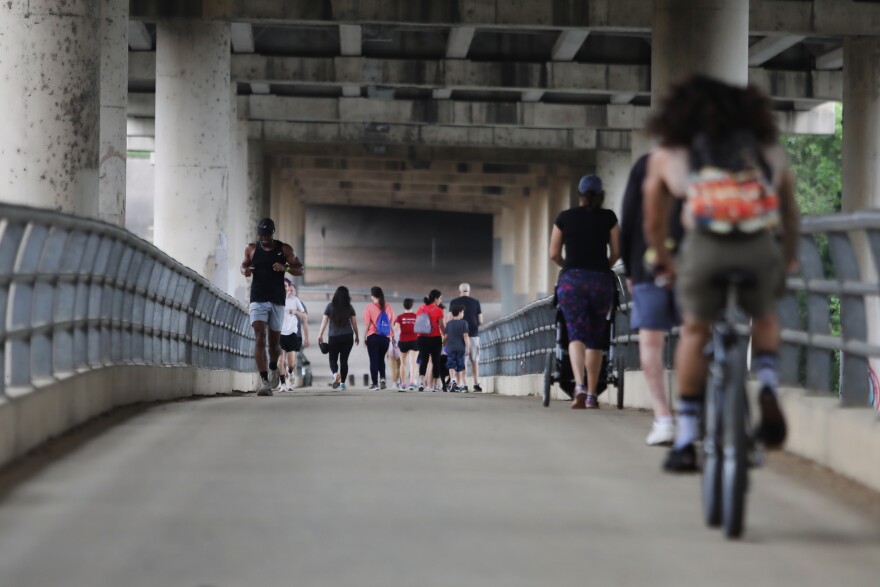 Runners, walkers and cyclists on the MoPac pedestrian bridge in March 2020. Above them is the bottom of the car bridge. On either side are grey, steel railings. One woman is pushing a baby stroller. Most people are wearing shorts and t-shirts.