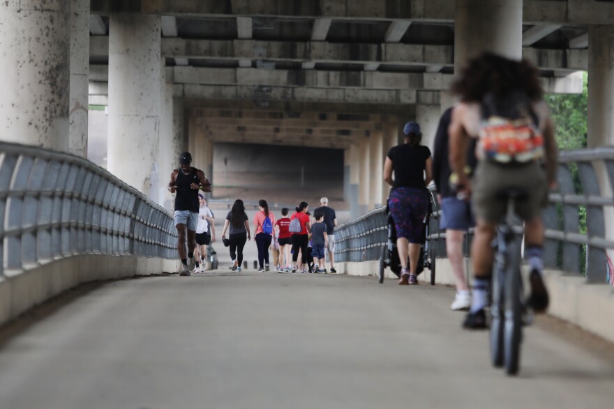 Runners, walkers and cyclists on the MoPac pedestrian bridge in March 2020. Above them is the bottom of the car bridge. On either side are grey, steel railings. One woman is pushing a baby stroller. Most people are wearing shorts and t-shirts. 