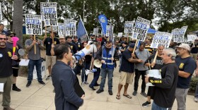 A crowd of people stand around a man in a suit with a megaphone. Many are holding signs that say, "CSU unfair to labor, Pay up or strike out."
