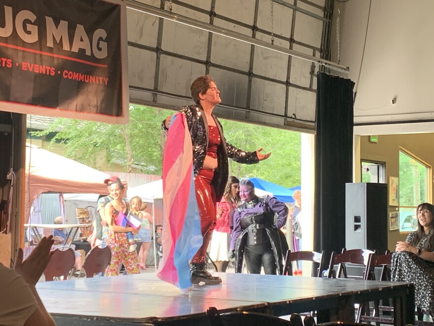  Blue Taboo, a drag king, performs on stage. They wear a tight, shiny red outfit with a sparkly black jacket and hold a  trans flag.