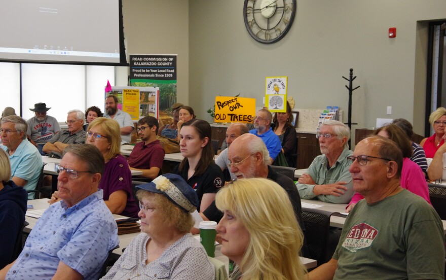 Rows of people sit at white tables, some even line the sides of the room in extra chairs. They all look to the left of the image, towards something off camera. Near the back, two signs can be seen being held by two attendees. One is a light orange sign that reads "Respect our old trees," the other is a yellow sign with a black and white depiction of a tree, above the image is a picture of an orange, furry creature that stands on two legs and has a large, yellow bushy mustache. It's the Lorax, and letters above him read "I speak for the trees." A sign to the back right labels the room as part of the Road Commission of Kalamazoo County. 