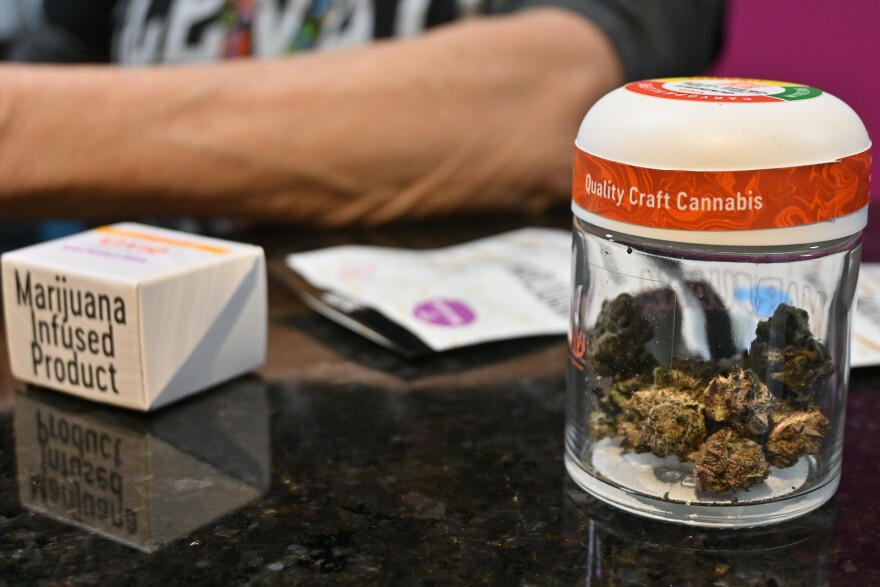 A small white box read "Marijuana Infused Product, sits next to a small white packet and a glass jar that contains marijuana flower inside. It is labeled "Quality Craft Cannabis."