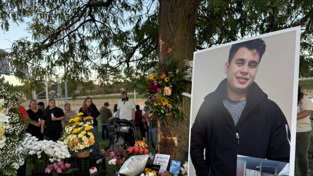 A poster-sized photo of a smiling man in a dark zip up sweater sits on an easel in front of a treet surrounded by colorful flowers. On the ground, white tea lights are arranged in the shape of a cross. 
