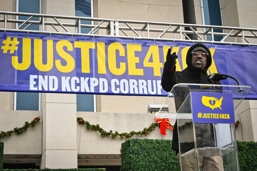 A man speaking at a podium gestures. He is standing in front of a sign that reads #Justice4KCK, End KCKPD Corruption."